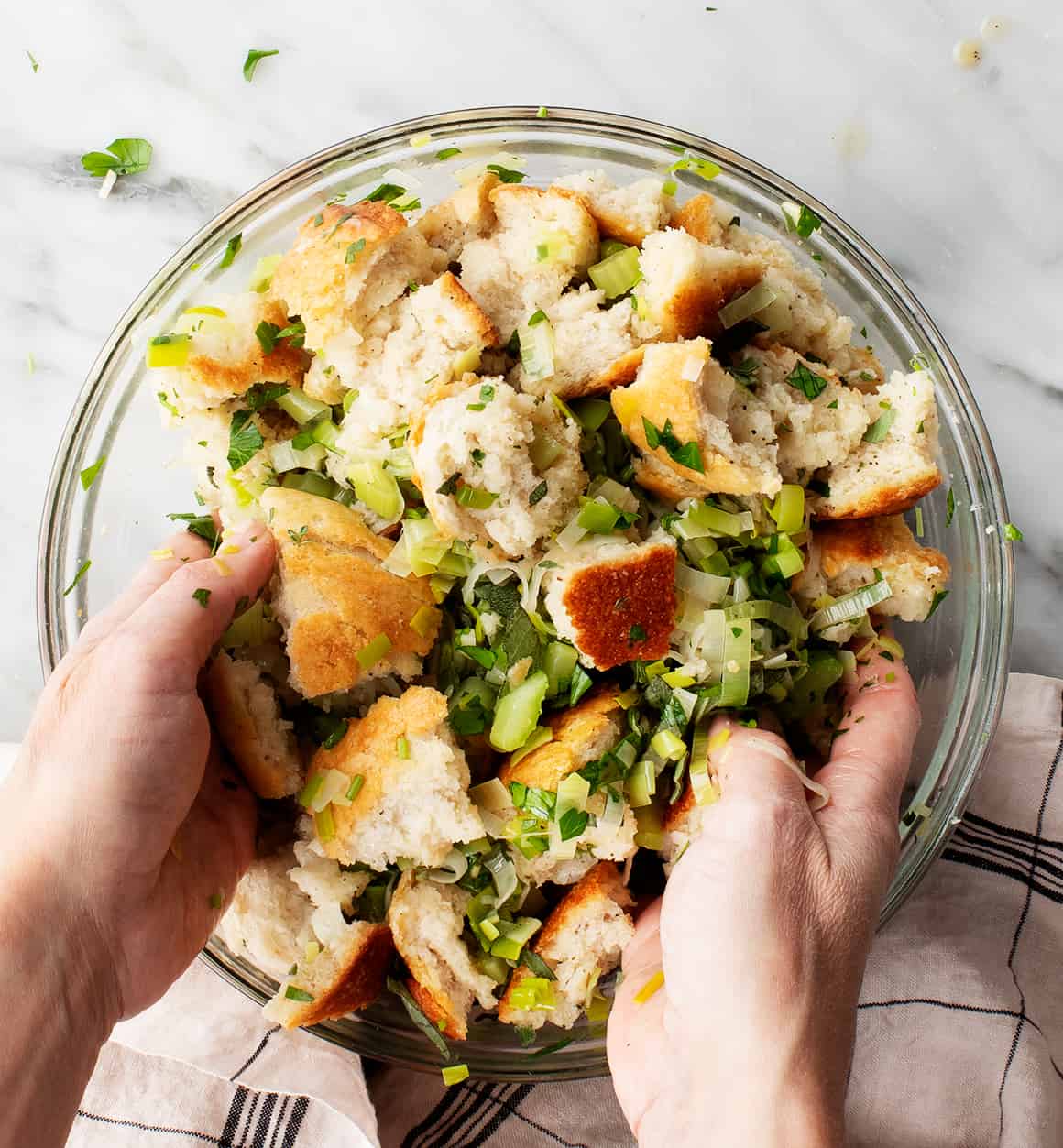 Hands mixing bread cubes with herbs, leeks, and celery